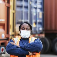 A mature African-American woman in her 40s working at a shipping port, cargo containers on trailers out of focus behind her. She is wearing a protective face mask, working during COVID-19, trying to prevent the spread of coronavirus. She is looking at the camera, arms folded.
